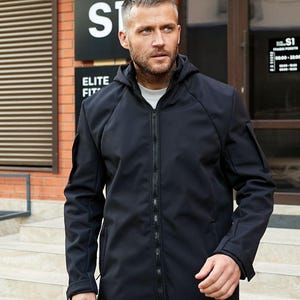Puede incluir: Una chaqueta softshell negra con cierre de cremallera y capucha. La chaqueta tiene un dobladillo recto y dos bolsillos en la parte delantera.