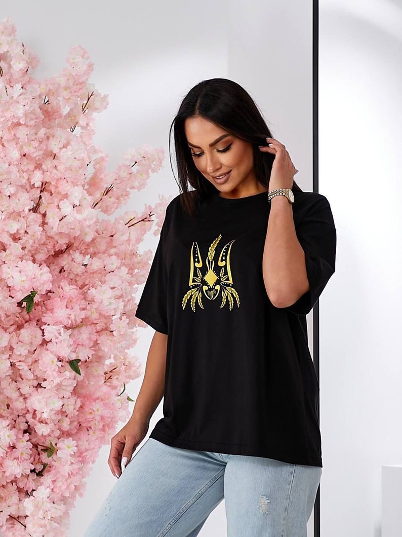 Puede incluir: Camiseta negra de gran tama&ntilde;o con un s&iacute;mbolo de tridente ucraniano bordado en oro.