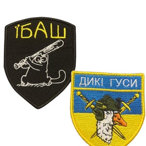 May include: Two embroidered shield-shaped patches. The black patch features a white cartoon cat holding a baseball bat and the yellow text "1БАШ". The blue and yellow patch depicts a goose wearing a green beret over crossed swords with the text "ДИКІ ГУСИ".