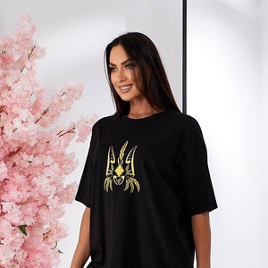 Puede incluir: Camiseta negra con un s&iacute;mbolo de tridente ucraniano bordado en oro.