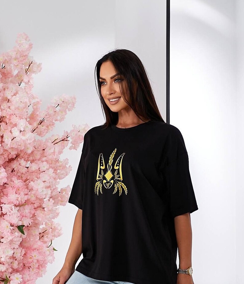 Puede incluir: Camiseta negra con un s&iacute;mbolo de tridente ucraniano bordado en oro.