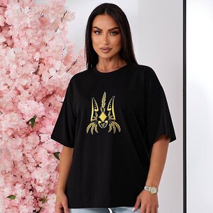 Puede incluir: Camiseta negra con un s&iacute;mbolo de tridente ucraniano bordado en oro.