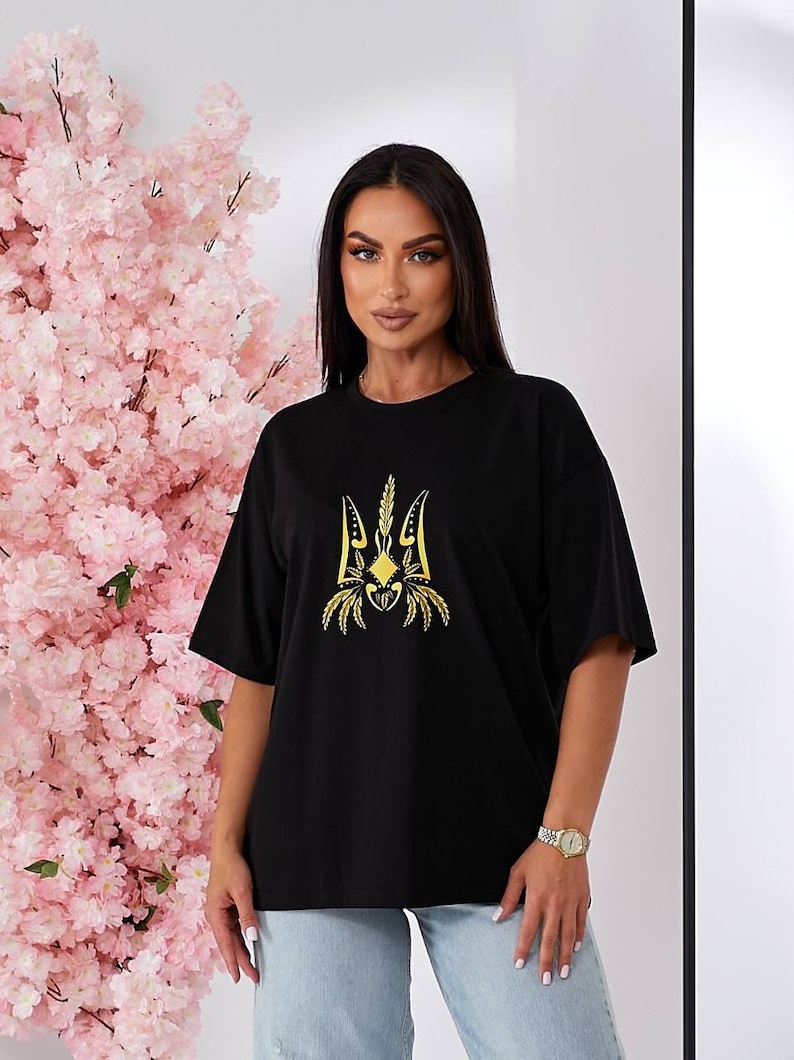 Puede incluir: Camiseta negra con un s&iacute;mbolo de tridente ucraniano bordado en oro.