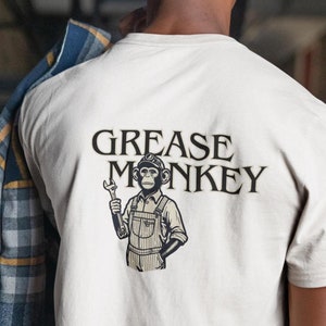 May include: A light beige t-shirt featuring the words "GREASE MONKEY" above a cartoon monkey wearing a cap and overalls, holding a wrench. The shirt is made of a soft material.