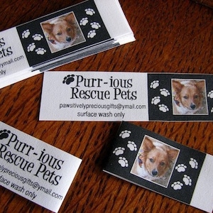 K&ouml;nnte beinhalten: Ein wei&szlig;er rechteckiger Aufkleber mit schwarzem Text, der "Purr-ious Rescue Pets" und die E-Mail-Adresse "pawsitivelypreciousgifts@ymail.com" lautet. Der Text "surface wash only" ist ebenfalls enthalten. Der Aufkleber hat einen schwarzen Rand und Pfotenabdr&uuml;cke an den R&auml;ndern. Ein Foto eines Hundes befindet sich in der Mitte des Aufklebers.