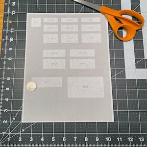 Pu&ograve; includere: Un foglio di carta bianco con varie scatole rettangolari di diverse dimensioni stampate su di esso. Le scatole sono etichettate con misure in pollici. Un paio di forbici con manici arancioni e una moneta d'argento si trovano su un tappetino da taglio nero.