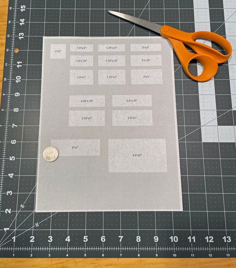 K&ouml;nnte beinhalten: Ein wei&szlig;es Blatt Papier mit verschiedenen rechteckigen K&auml;sten, die darauf gedruckt sind. Die K&auml;sten sind mit Ma&szlig;en in Zoll beschriftet. Eine Schere mit orangefarbenen Griffen und eine Silberm&uuml;nze liegen auf einer schwarzen Schneidematte.
