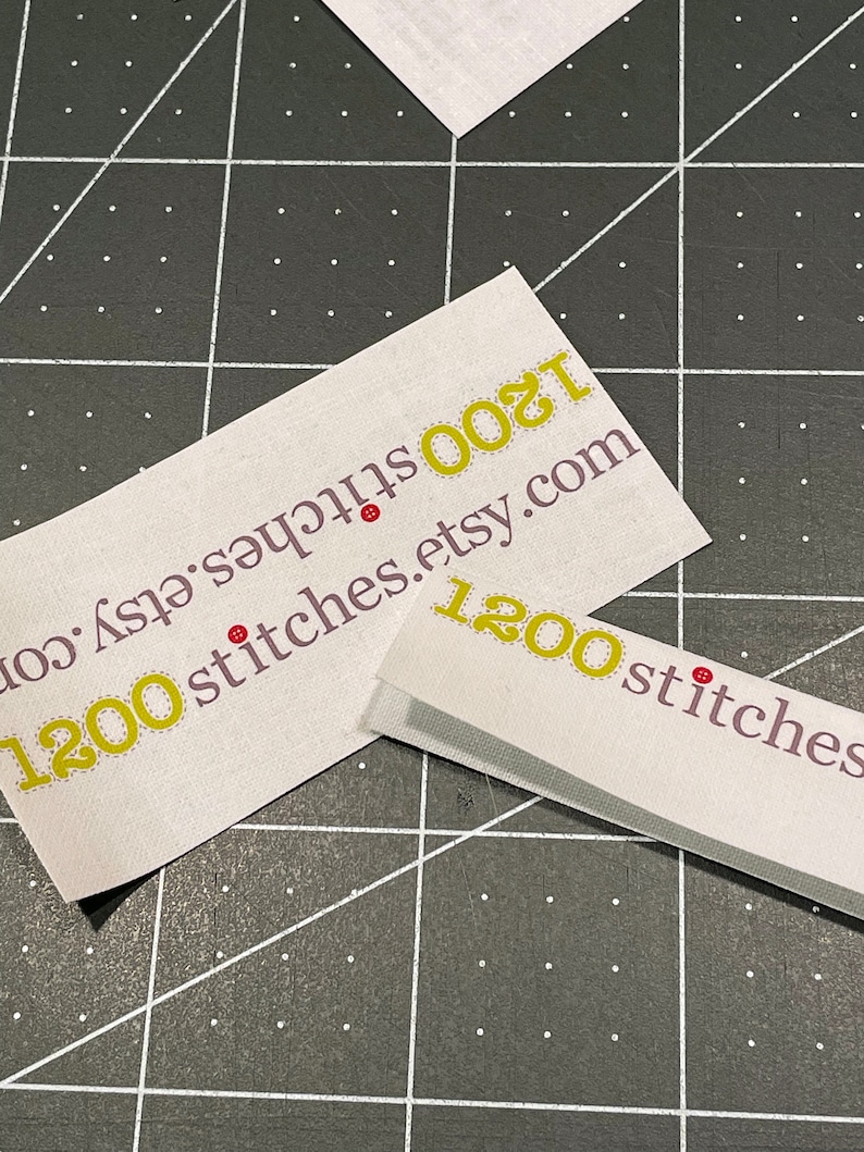 K&ouml;nnte beinhalten: Zwei wei&szlig;e Etiketten mit gelbem Text und roten Punkten. Der Text auf den Etiketten lautet "1200 stitches.etsy.com".
