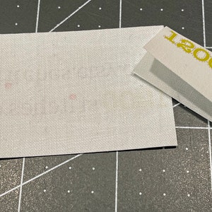 K&ouml;nnte beinhalten: Ein wei&szlig;es Stoffquadrat mit einigen kleinen rosa Punkten. Der Stoff liegt auf einer schwarz-wei&szlig;en Gitteroberfl&auml;che. Ein wei&szlig;es Papier mit dem Text "1200" in Gelb ist teilweise im oberen rechten Eck des Bildes sichtbar.