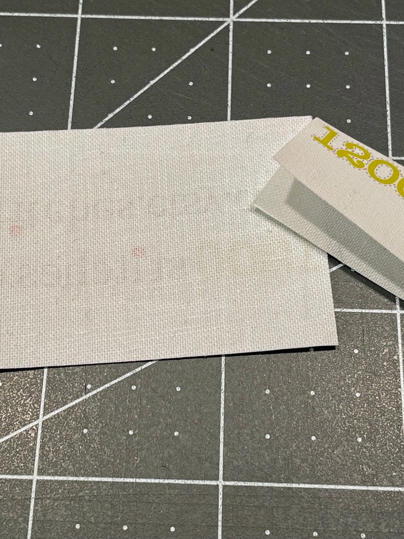 K&ouml;nnte beinhalten: Ein wei&szlig;es Stoffquadrat mit einigen kleinen rosa Punkten. Der Stoff liegt auf einer schwarz-wei&szlig;en Gitteroberfl&auml;che. Ein wei&szlig;es Papier mit dem Text "1200" in Gelb ist teilweise im oberen rechten Eck des Bildes sichtbar.