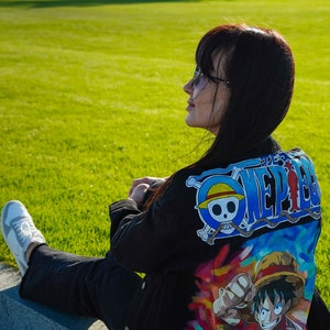 K&ouml;nnte beinhalten: Schwarze Jeansjacke mit einem farbenfrohen One Piece Anime-Motiv auf dem R&uuml;cken. Das Design beinhaltet ein blau-wei&szlig;es Logo. Die Person tr&auml;gt eine schwarze Hose und wei&szlig;e Turnschuhe. Die Szene spielt im Freien.