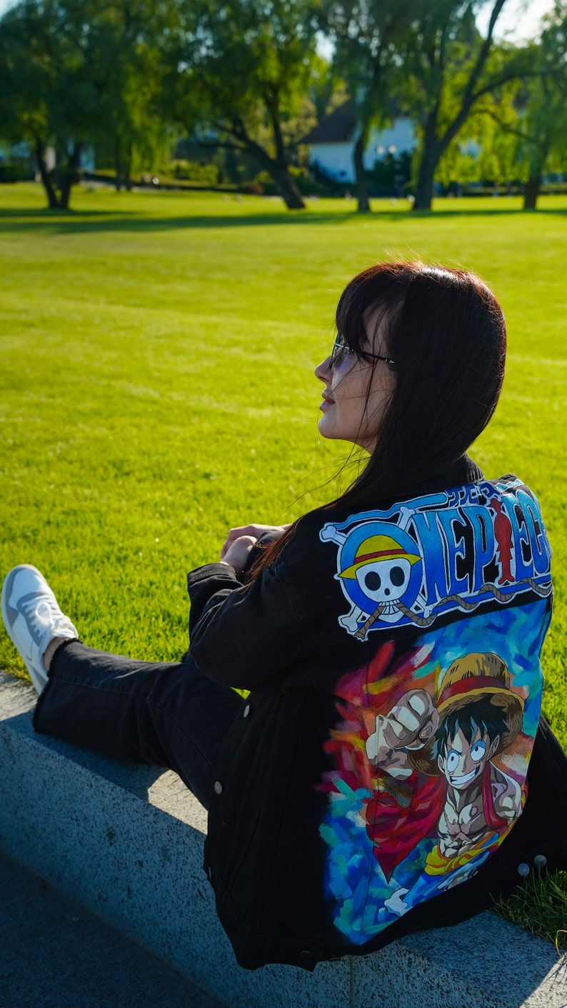 K&ouml;nnte beinhalten: Schwarze Jeansjacke mit einem farbenfrohen One Piece Anime-Motiv auf dem R&uuml;cken. Das Design beinhaltet ein blau-wei&szlig;es Logo. Die Person tr&auml;gt eine schwarze Hose und wei&szlig;e Turnschuhe. Die Szene spielt im Freien.