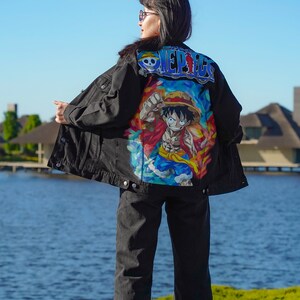 K&ouml;nnte beinhalten: Schwarze Jeansjacke mit einem farbenfrohen Cartoon-Motiv auf dem R&uuml;cken. Auf der Jacke steht in Blau und Wei&szlig; das Wort "ONE PIECE". Die Person tr&auml;gt eine schwarze Jeans und wei&szlig;e Turnschuhe.