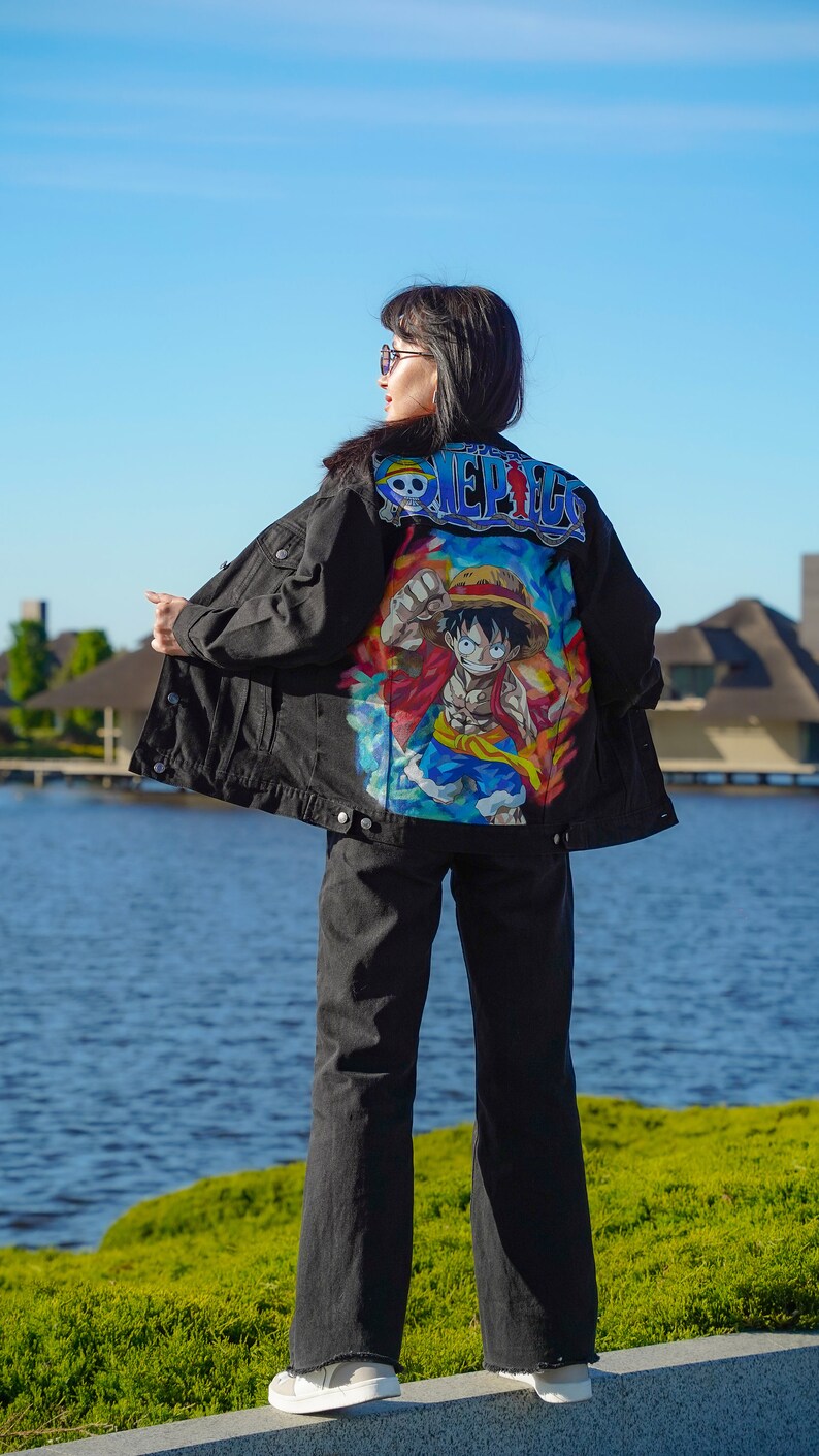 K&ouml;nnte beinhalten: Schwarze Jeansjacke mit einem farbenfrohen Cartoon-Motiv auf dem R&uuml;cken. Auf der Jacke steht in Blau und Wei&szlig; das Wort "ONE PIECE". Die Person tr&auml;gt eine schwarze Jeans und wei&szlig;e Turnschuhe.
