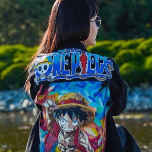 K&ouml;nnte beinhalten: Schwarze Jeansjacke mit einer farbenfrohen Grafik einer Zeichentrickfigur. Die Jacke tr&auml;gt das Wort "ONE PIECE" in blauen und wei&szlig;en Buchstaben &uuml;ber den Schultern. Das Kunstwerk zeigt eine Figur mit Strohhut.