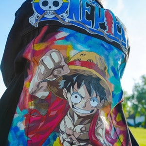 K&ouml;nnte beinhalten: Schwarze Jeansjacke mit farbenfrohem Cartoon-Motiv. Die Jacke hat den Schriftzug "ONE PIECE" in Blau und Wei&szlig; auf den Schultern und eine gro&szlig;e Illustration auf dem R&uuml;cken.