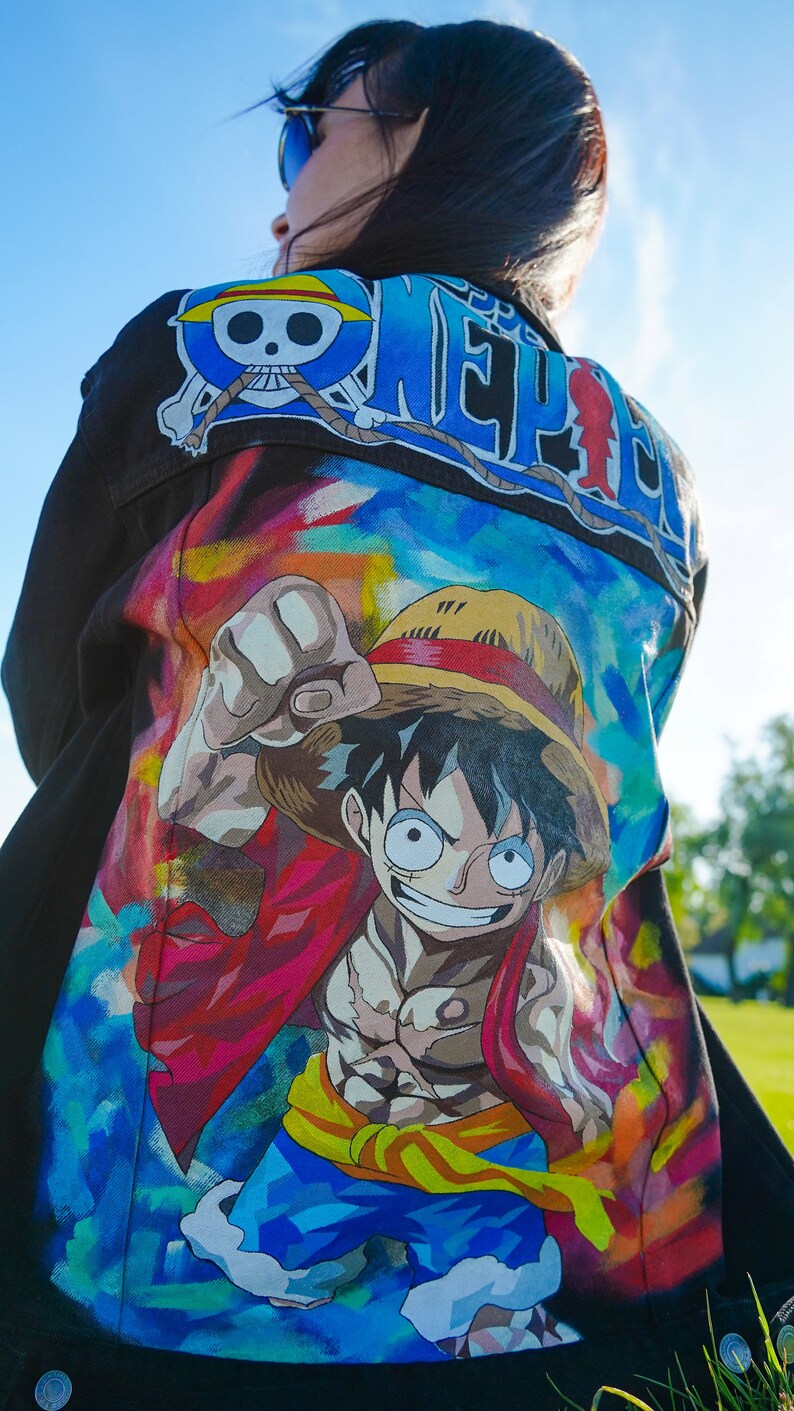 K&ouml;nnte beinhalten: Schwarze Jeansjacke mit farbenfrohem Cartoon-Motiv. Die Jacke hat den Schriftzug "ONE PIECE" in Blau und Wei&szlig; auf den Schultern und eine gro&szlig;e Illustration auf dem R&uuml;cken.