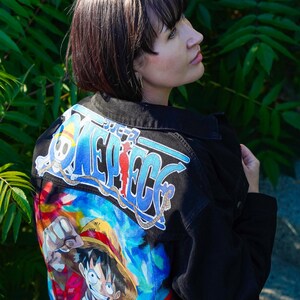 K&ouml;nnte beinhalten: Schwarze Jeansjacke mit einer farbenfrohen Grafik eines Charakters aus der Anime-Serie One Piece. Auf der R&uuml;ckseite der Jacke befindet sich das Serienlogo in Blau und Wei&szlig; mit einer lebendigen Illustration darunter.