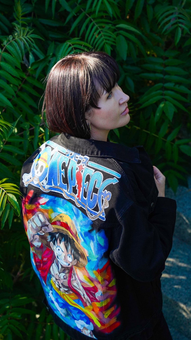 K&ouml;nnte beinhalten: Schwarze Jeansjacke mit einer farbenfrohen Grafik eines Charakters aus der Anime-Serie One Piece. Auf der R&uuml;ckseite der Jacke befindet sich das Serienlogo in Blau und Wei&szlig; mit einer lebendigen Illustration darunter.