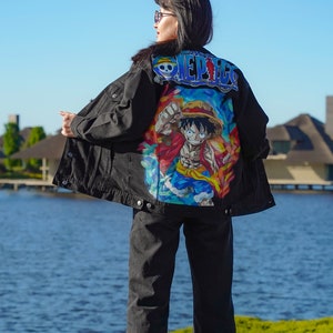 K&ouml;nnte beinhalten: Schwarze Jeansjacke mit einem farbenfrohen Cartoon-Motiv und dem Schriftzug "ONE PIECE" auf dem R&uuml;cken. Die Jacke wird mit einer schwarzen Jeans und wei&szlig;en Turnschuhen kombiniert. Im Hintergrund ein See und blauer Himmel.