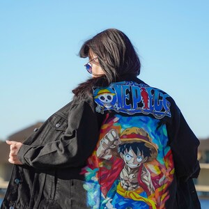 K&ouml;nnte beinhalten: Schwarze Jeansjacke mit farbenfroher Grafik eines Charakters aus der Anime-Serie One Piece. Auf der R&uuml;ckseite der Jacke steht das Wort "ONE PIECE" in Blau und Wei&szlig;. Die Person tr&auml;gt eine schwarze Hose.