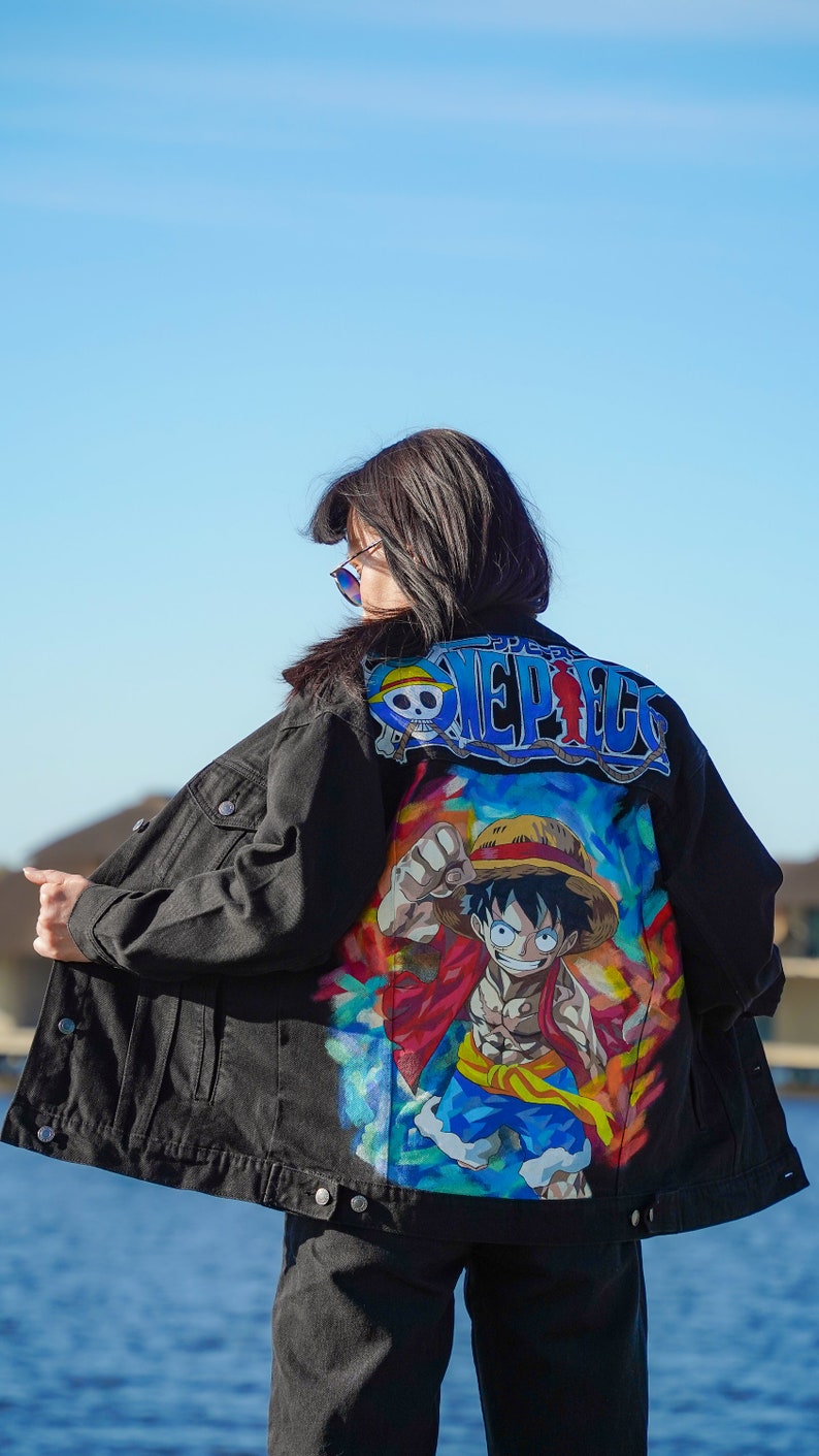 K&ouml;nnte beinhalten: Schwarze Jeansjacke mit farbenfroher Grafik eines Charakters aus der Anime-Serie One Piece. Auf der R&uuml;ckseite der Jacke steht das Wort "ONE PIECE" in Blau und Wei&szlig;. Die Person tr&auml;gt eine schwarze Hose.