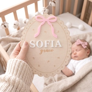May include: A wooden sign with the name "SOFIA grace" in white and gold lettering, topped with a pink bow. The sign has a scalloped edge and floral accents. The sign is held near a sleeping baby in a crib.