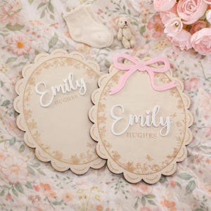 以下が含まれることがあります： 白い筆記体で「Emily」という名前が書かれた、スカラップエッジの2つの木製楕円形プラーク。1つのプラークにはピンクのリボンが付いています。どちらにも花の模様があり、「Emily」の下に「Hughes」という名前があります。