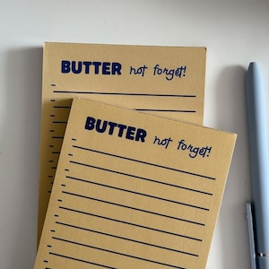 May include: Two tan notepads with the text "BUTTER not forget!" in blue script. The notepads have horizontal lines and a vertical ruler on the left side. A light blue pen is on the right side of the notepads.