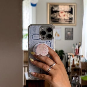 May include: A clear phone case with a light blue patterned design and a pink seashell phone grip. The phone case is held in front of a mirror, reflecting the room's interior. The person's hand is visible, adorned with rings and yellow nail polish.