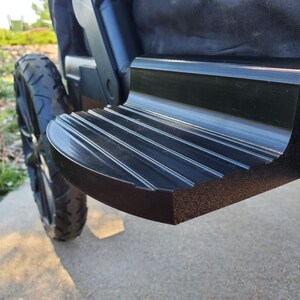 May include: A black, semi-circular step attachment with a ribbed, non-slip surface texture. The component is mounted onto the horizontal frame of a collapsible wagon, positioned directly in front of a large, black, treaded rubber wheel.
