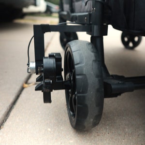 May include: Close-up of a black wagon wheel with a tread pattern. The wheel is attached to a black metal frame with a gear mechanism.