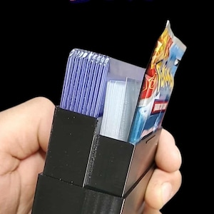 May include: Black portable TCG box with the words "PORTABLE TCG BOX" in colorful letters. The box is open, revealing stacks of trading cards and a sealed pack. The box is held in a hand against a black background.