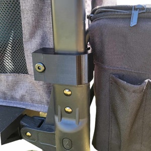 May include: A black metal frame with a black fabric bag attached. The bag has a zipper and a pocket. The frame has a black plastic handle and a black plastic wheel.