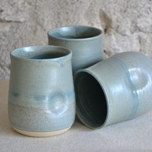 May include: Three ceramic tumblers with a smooth, matte, sage-green glaze. Each vessel features a subtle, circular thumb indentation on the side and a contrasting, unglazed beige base. The cups have a slightly flared rim and a cylindrical shape.
