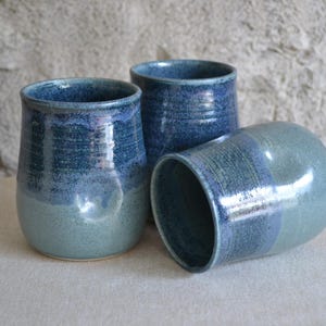 May include: Three ceramic tumblers with a glossy finish, featuring a two-tone colour scheme of deep navy blue at the top and a lighter, muted teal-blue at the base. The vessels have a rounded, cylindrical shape with a smooth, reflective surface.