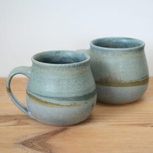 May include: Two handmade ceramic mugs. The mugs are a light blue-green colour with a speckled texture and a horizontal band of olive green. They have rounded bodies, sturdy handles, and sit on a wooden surface.