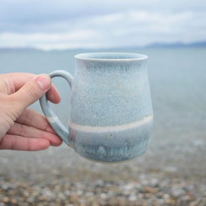 May include: A ceramic mug with a rounded body and a curved handle, featuring a mottled light blue and grey glaze with a distinct horizontal white band encircling the middle section of the vessel, showcasing a smooth, glossy finish throughout.