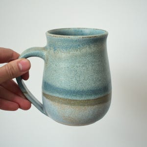 May include: A handmade ceramic mug with a large handle. The mug features a speckled, textured surface with bands of blue, tan, and grey. The mug is held by a hand against a white background. The mug is approximately 13 cm tall.