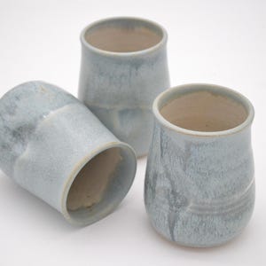 May include: Three ceramic tumblers with a light blue and grey mottled glaze. Each vessel features a rounded, slightly tapered body with a smooth rim and a visible cream-coloured interior. The finish shows subtle variations in colour and texture.