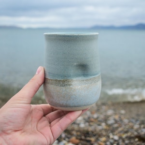 May include: A ceramic tumbler featuring a smooth, matte finish with a gradient colour scheme transitioning from a soft, muted blue at the top to a sandy, beige-toned base. The vessel has a slightly curved, cylindrical shape with a subtle indentation.