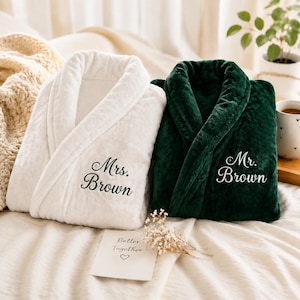 Puede incluir: Una bata de felpa blanca bordada con el texto Mrs. Brown y una bata de felpa verde oscuro bordada con el texto Mr. Brown. Ambas batas cuentan con cuello de chal y textura suave, presentadas una al lado de la otra sobre una superficie neutra.
