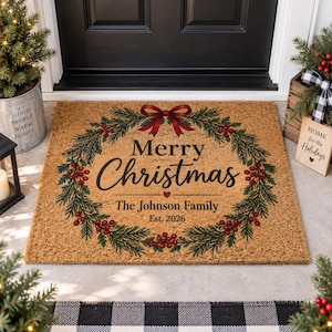 Puede incluir: Un felpudo rectangular de fibra de coco color canela con una corona de pino verde impresa, bayas rojas y un lazo rojo. El texto dice Merry Christmas, The Johnson Family y Est. 2026 en letras negras con un pequeño corazón rojo en el centro.
