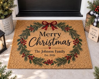 Puede incluir: Un felpudo rectangular de fibra de coco color canela con una corona de pino verde impresa, bayas rojas y un lazo rojo. El texto dice Merry Christmas, The Johnson Family y Est. 2026 en letras negras con un pequeño corazón rojo en el centro.