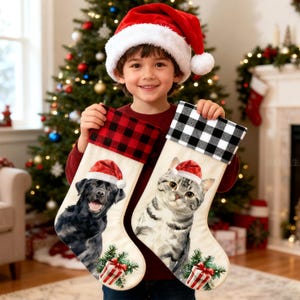 May include: Two Christmas stockings with festive designs. One features a black Labrador wearing a Santa hat, the other a gray tabby cat in a Santa hat. Both stockings have a cream-colored base, a gift, and a green pine tree.