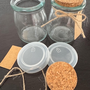 May include: Two clear glass jars with rounded bodies and wide openings. One jar has a cork lid, tied with twine and a blank tag. Two clear plastic lids and a cork lid are also present, along with a length of twine and a blank tag.