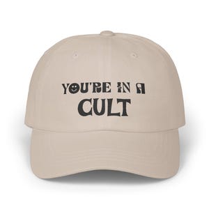 May include: A beige baseball cap with the phrase "YOU'RE IN A CULT" printed in black. The word "YOU'RE" has a smiley face replacing the "O". The cap has a curved brim and a rounded crown.