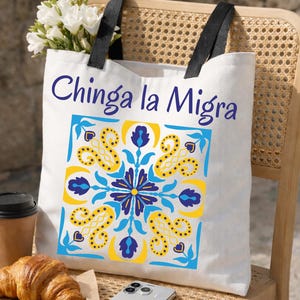 May include: White tote bag with black handles featuring the text "Chinga la Migra" in blue script above a square design with yellow, blue, and white floral patterns. The bag is on a wicker chair with a croissant, coffee, and a phone.