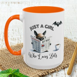 May include: White ceramic mug with an orange interior and handle. The mug features the text "JUST A GIRL Who Loves Bats" and an illustration of a bat reading a newspaper. A small bat silhouette is above the text. The mug is 15 oz.