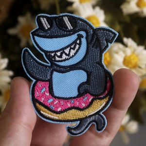 May include: A blue shark wearing sunglasses and holding a pink and white doughnut. The shark is embroidered on a patch.